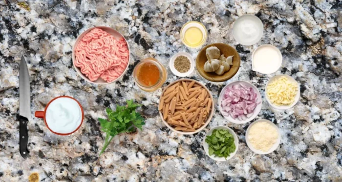 Ingredients for one-skillet cheesy ground chicken pasta laid out on a granite countertop, including ground chicken, pasta, shredded cheese, garlic, onions, parsley,  Parmesan cheese, milk, olive oil, chicken stock, oregano, and salt.