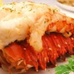 Close-up of a butter-baked lobster tail with golden, tender meat on a white plate, garnished with fresh parsley.