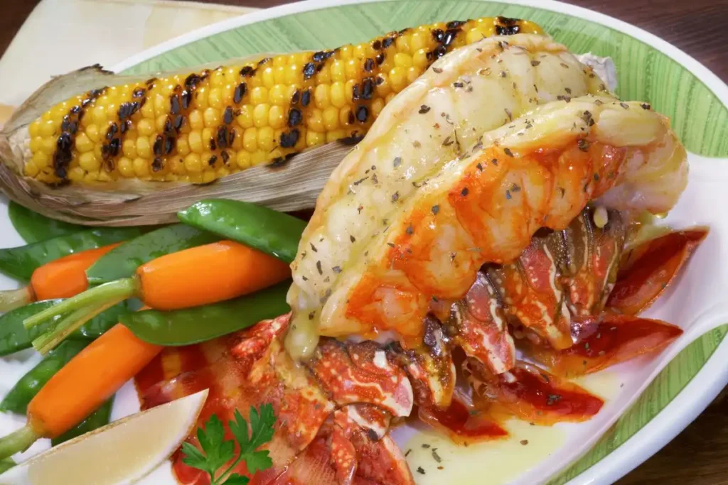 Butter-basted baked lobster tail served with grilled corn, baby carrots, and sugar snap peas on a white plate.